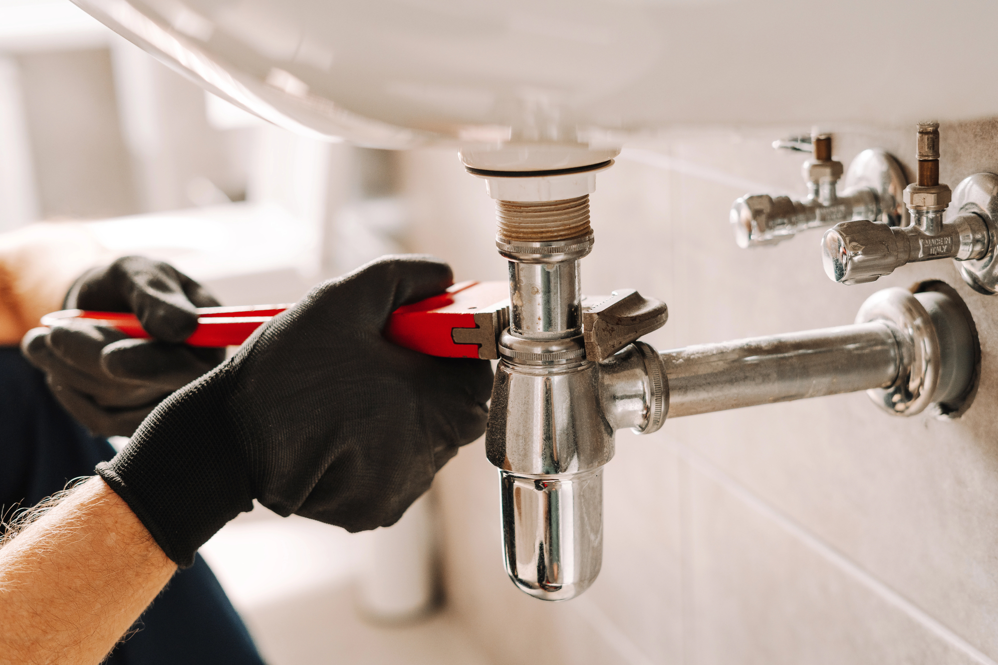 A person wearing black gloves uses a red wrench to tighten or loosen plumbing pipes under a bathroom sink.
