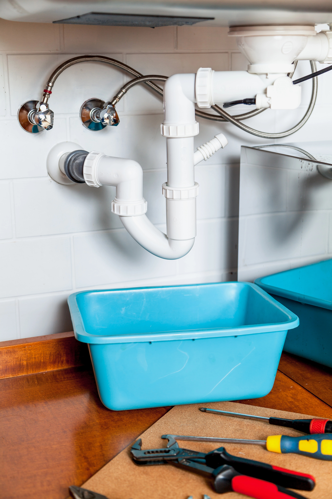 White plastic plumbing pipes under a sink with a blue basin positioned below and various plumbing tools on the wooden surface nearby.