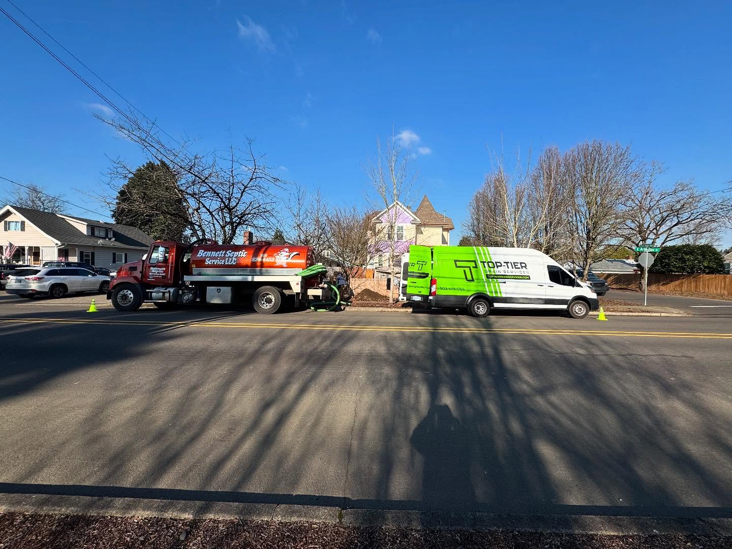 Two work vehicles, a red truck and a green-and-white van, are parked on a suburban street near a house with leafless trees and blue skies overhead.