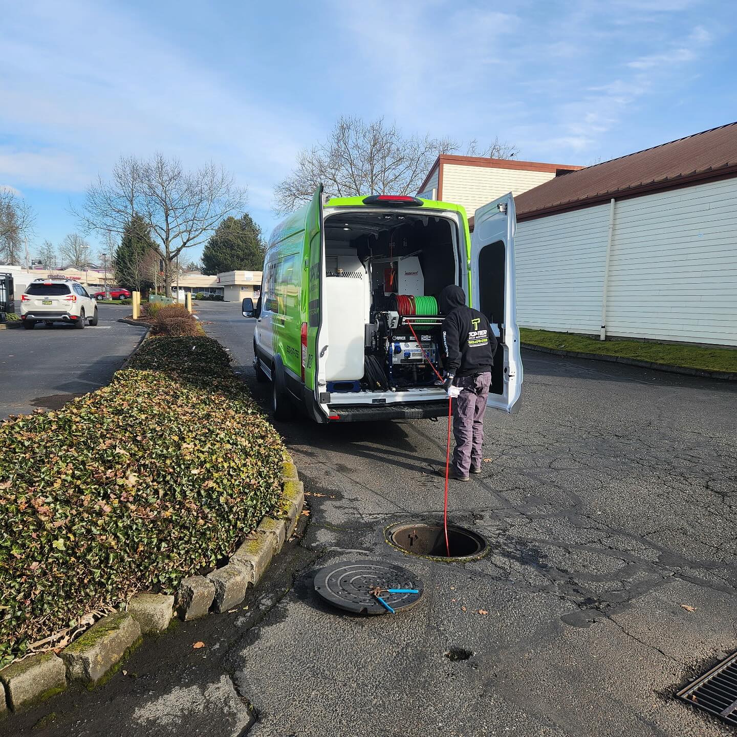 A person stands at the back of a green utility van, using equipment connected to an open manhole in a parking lot.