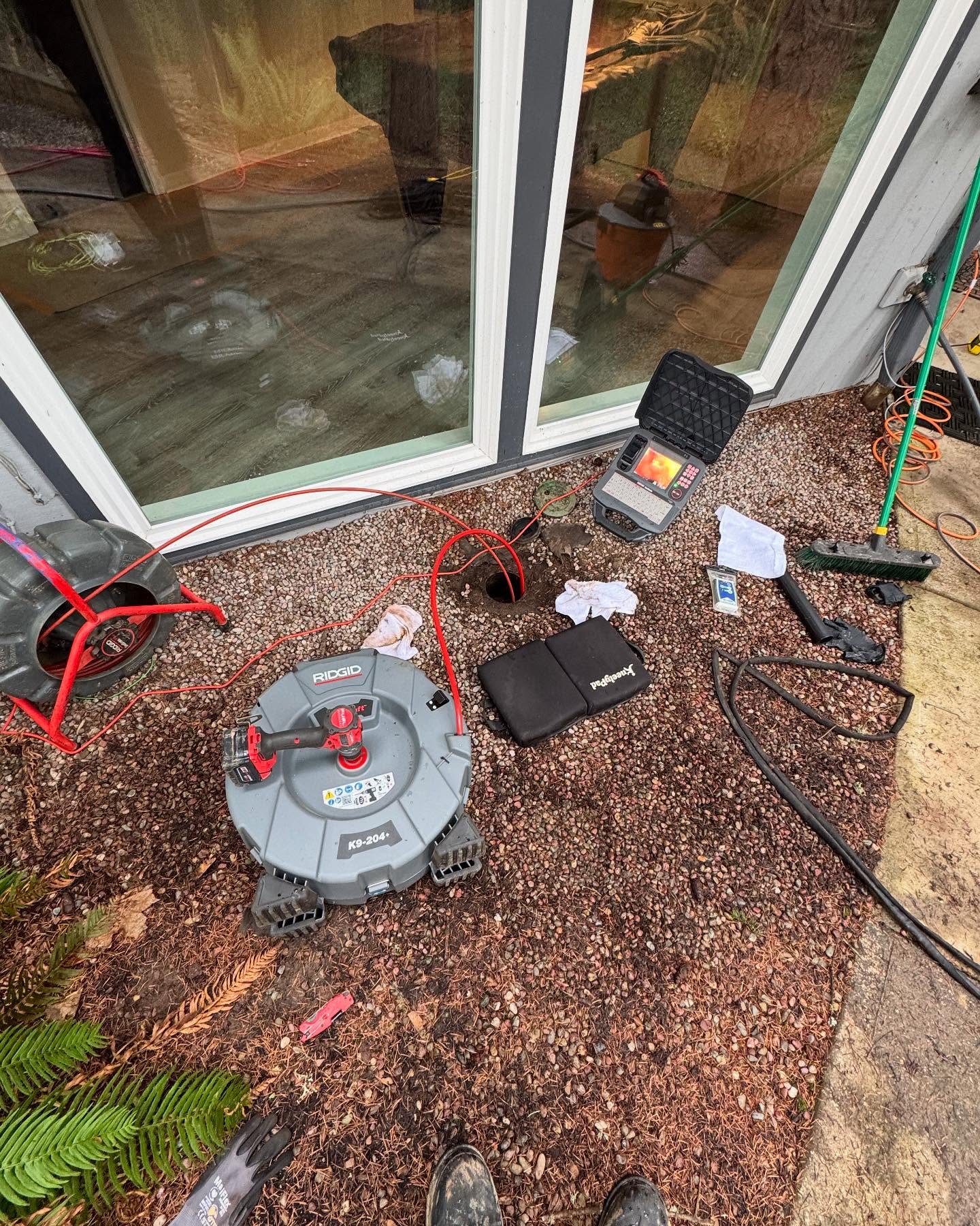 Plumbing inspection equipment, including a Ridgid camera reel and monitor, is set up outside a house near a glass door on gravel, with cables and tools scattered around.